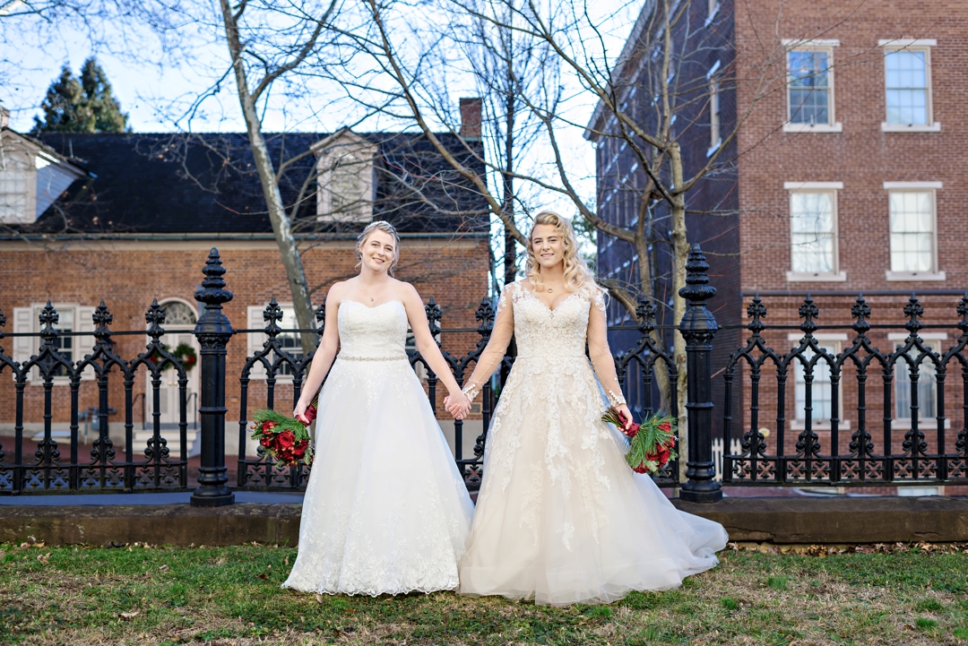 Two brides holding red roses stand in their wedding dresses before their wedding ceremony.