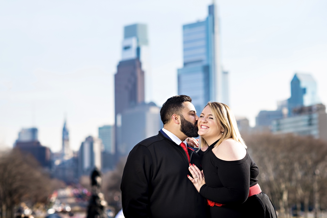 A man kisses his fiance on the steps of the art museum with the Philly skyline in the background.