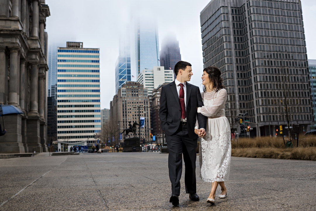 A bride and groom walk next to city hall in Philadelphia on a foggy day.