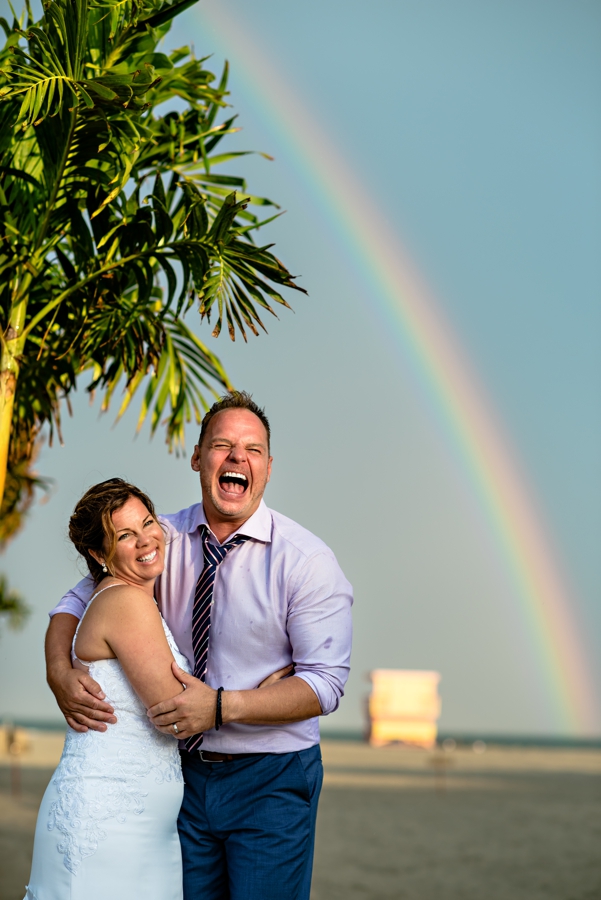 A newly married couple laugh on the beach with a rainbow in the sky behind them in Cape May.