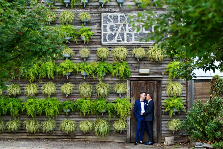 A same sex couple hold each other in front a wall of green plants at their wedding at Terrain at Styers in Glen Mills, PA.