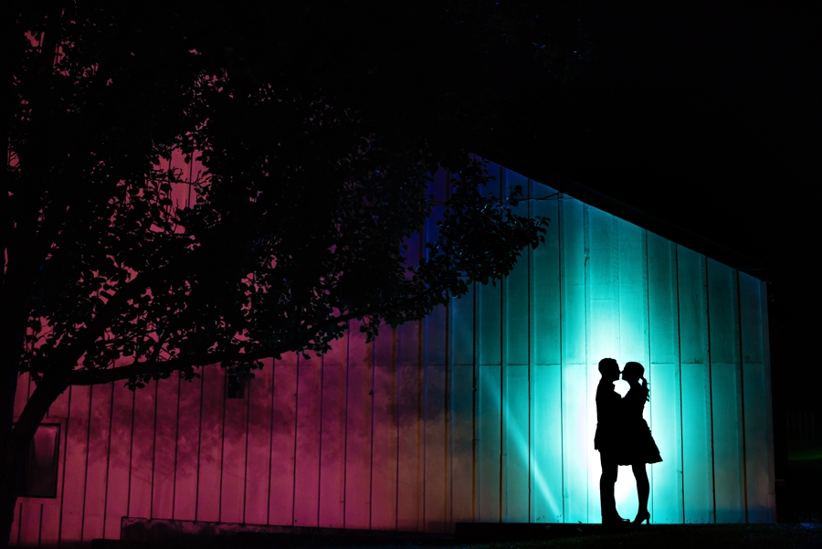 A silhouette of a wedding couple in front of a barn splashed with blue and pink color.