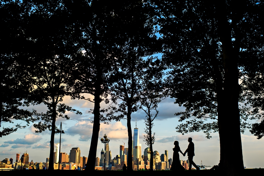 A bride to be and her fiance walk along the trees with the New York City Skyline in the background for their Hoboken Engagement Session.