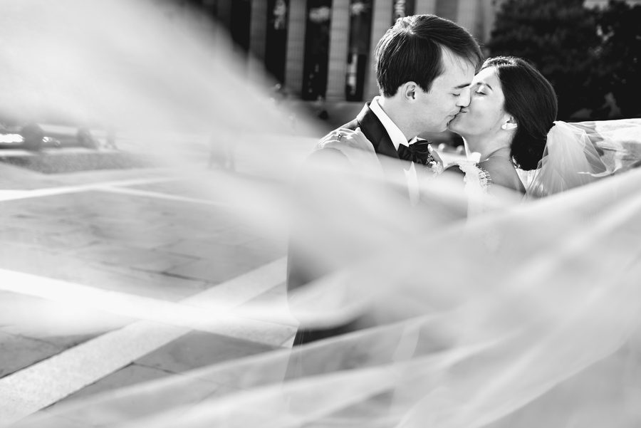 A bride and groom kiss on the Philadelphia Museum of Art steps with the brides full cathedral veil blowing in the wind during the Union League of Philadelphia Wedding.