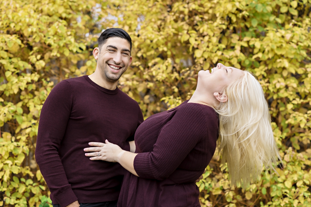 An engaged couple laugh in front of yellow fall leaves.