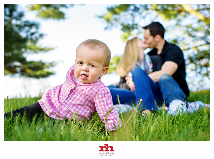 Portrait of a baby in the grass as her parents kiss in the background at Tyler State Park in Bucks County PA.