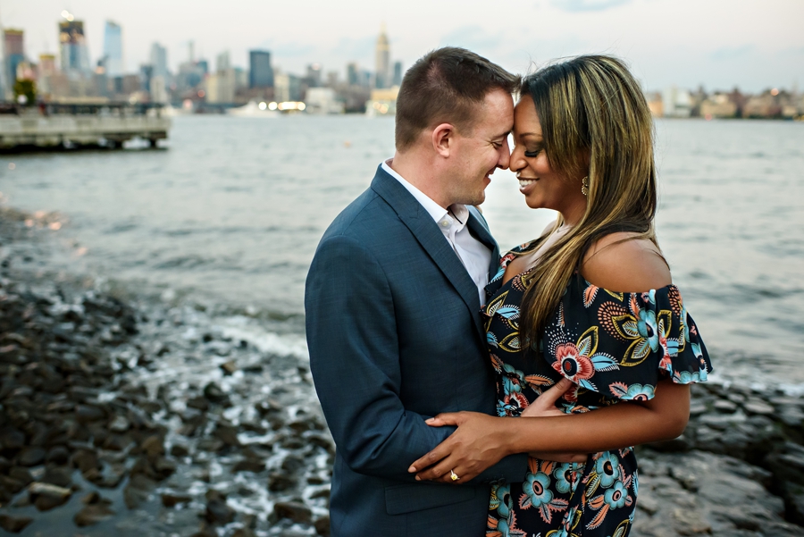 A man and woman hold each other on the rocks along the Hudson River in Hoboken with NYC in the background during their Engagement Session.