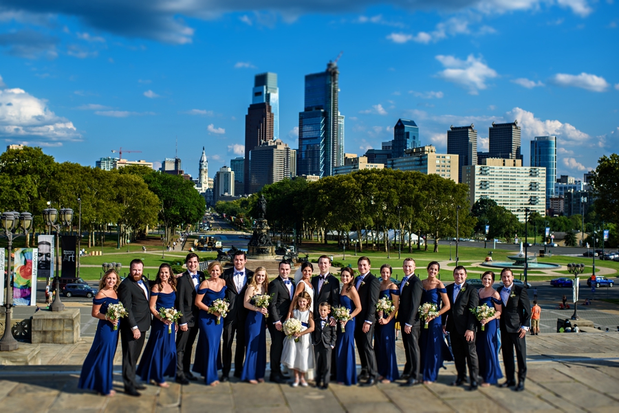 A large wedding party standing on the Philadelphia Art Museum steps with the Philly Skyline in the background during the Union League of Philadelphia Wedding.